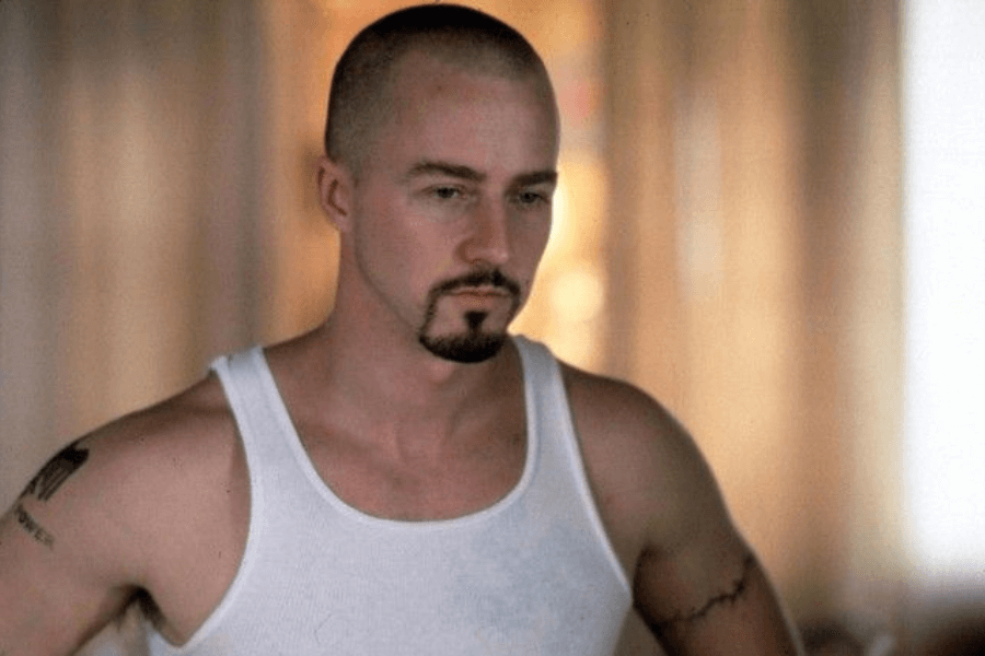 This image shows a man with a shaved head, a goatee, and tattoos on his arms, wearing a white tank top. His serious expression and stance convey intensity, with a softly lit indoor background creating a contemplative mood.