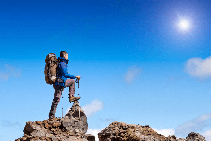 A hiker wearing outdoor gear and a backpack stands on a rocky peak, gazing into the distance under a bright blue sky with the sun shining. The scene conveys a sense of adventure and accomplishment.