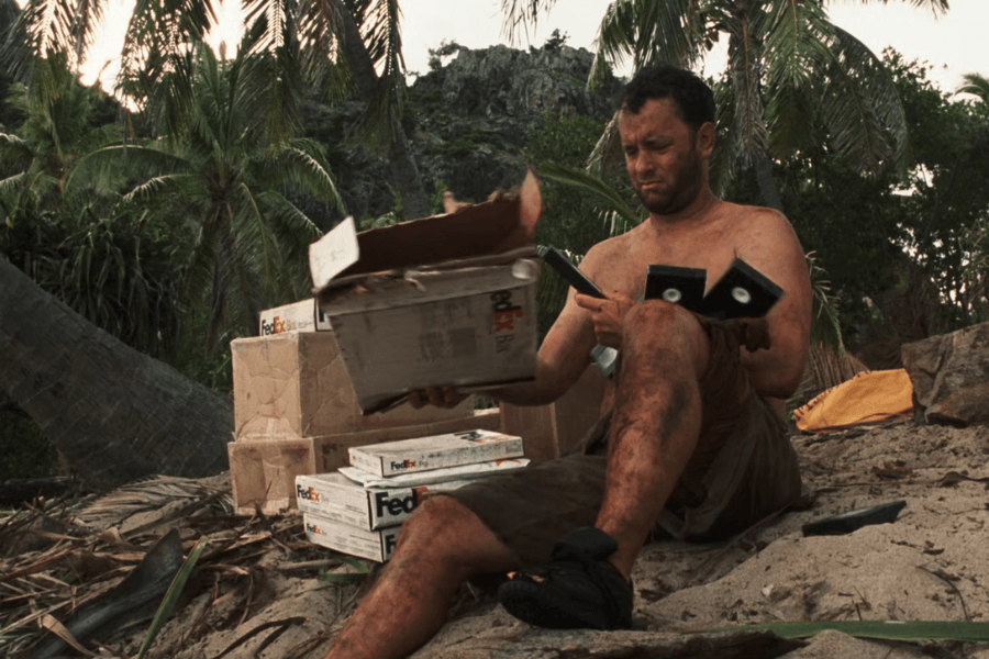 This image shows a shirtless man sitting on a sandy beach surrounded by FedEx packages, some opened, while examining their contents. The setting includes palm trees and rugged natural terrain, suggesting a survival scenario on a remote island. The man's appearance is rugged and dirty, reflecting the challenging environment.