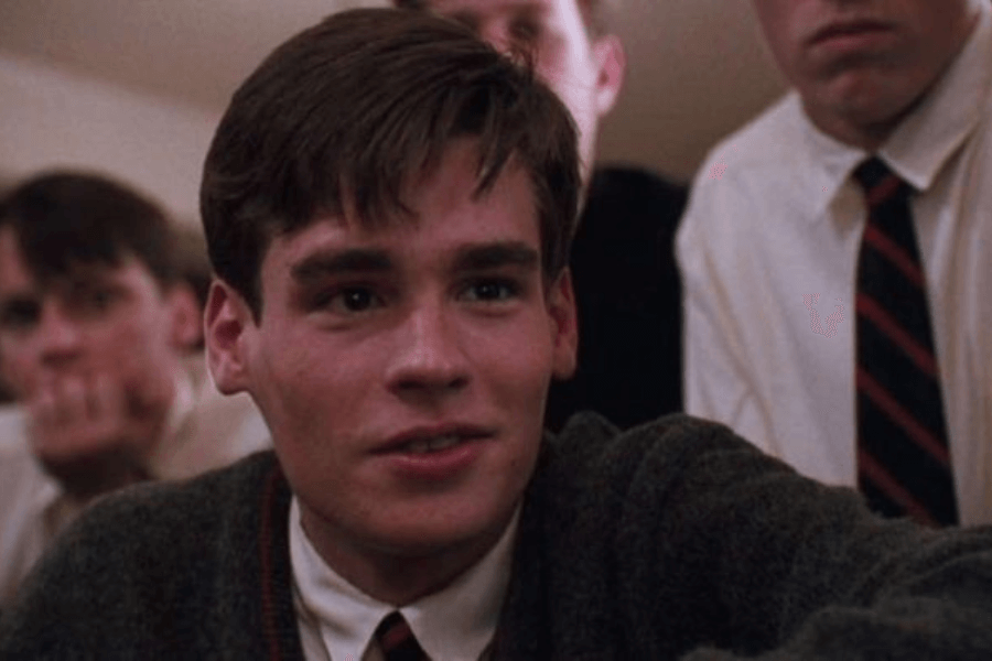 This image shows a young man with a warm expression, dressed in a white shirt, tie, and dark sweater, seated among a group of similarly dressed peers. The setting appears to be a formal or academic environment, with an air of camaraderie and attentiveness. His gaze and slight smile convey enthusiasm or engagement in the moment.