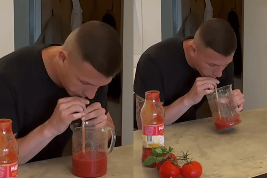 The image features a man sitting at a table attempting to drink a large quantity of tomato juice using a straw. On the table, there is a measuring jug filled with tomato juice, a bottle labeled with a tomato sauce brand, and fresh tomatoes with basil leaves for decoration. The setting suggests a challenge or record attempt involving speed or quantity, with the man focused on completing the task. The simple kitchen environment highlights the intensity of the activity.