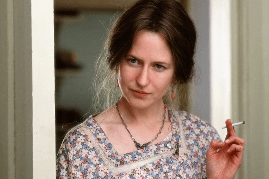 This image shows a woman with a thoughtful and slightly mischievous expression, wearing a floral-patterned dress and holding a cigarette. Her hair is styled loosely, and she stands in a softly lit indoor setting, framed by a doorway. The atmosphere suggests a period or intimate moment.