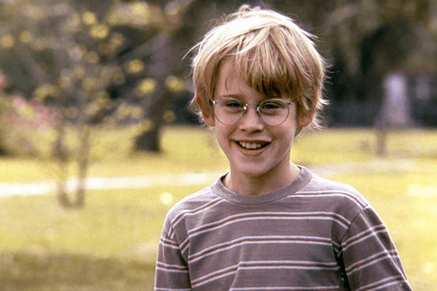 This image features a cheerful young boy with blonde hair and round glasses, smiling brightly while standing outdoors. He is wearing a striped shirt, and the background includes a sunny park with soft, blurred greenery and trees. The scene radiates warmth and innocence, capturing a carefree childhood moment.