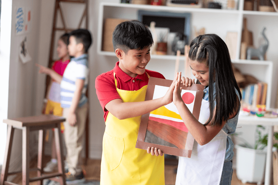 Two children wearing aprons share a high-five while holding up a colorful painting in a bright, creative classroom. Other kids and art supplies are visible in the background, emphasizing a joyful learning environment.