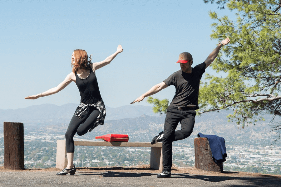 This image features two individuals dancing outdoors on a paved area with a scenic city and mountain view in the background. The woman on the left wears a black outfit with a plaid shirt tied around her waist, while the man on the right wears a black outfit and a cap. Their playful poses and the bright sunlight create a lighthearted and joyful atmosphere.