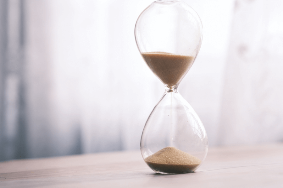 A glass hourglass with sand flowing from the top chamber to the bottom sits on a wooden surface. The soft, blurred background creates a serene and timeless atmosphere.