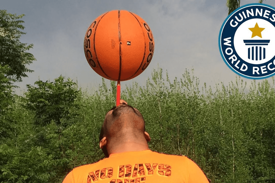 The image features a man balancing a basketball on a red toothbrush held in his mouth, showcasing an impressive display of skill and concentration. The background includes greenery and an open sky, creating a natural outdoor setting. A Guinness World Records logo is visible, indicating this is part of an official record attempt or achievement, emphasizing the unique and creative nature of the feat.