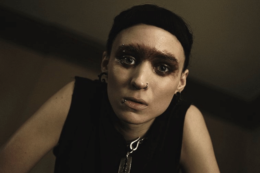 This image shows a woman with a striking and edgy appearance, featuring dark makeup, multiple piercings, and a bold, shaved eyebrow style. She wears a sleeveless black outfit and gazes intently at the camera in a dimly lit setting, creating a raw and intense atmosphere.