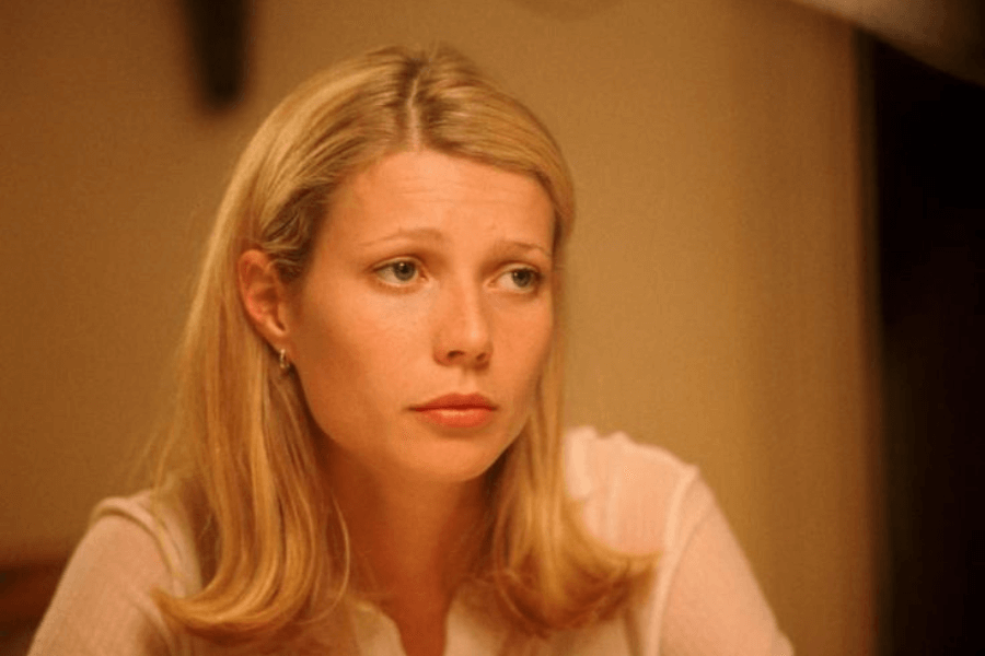 This image shows a woman with straight blonde hair, wearing a simple white blouse, seated indoors with a soft, warm light illuminating her face. Her expression is contemplative and slightly melancholic, suggesting she is deep in thought or processing an emotional moment. The subdued lighting and neutral background emphasize the intimate, reflective tone.