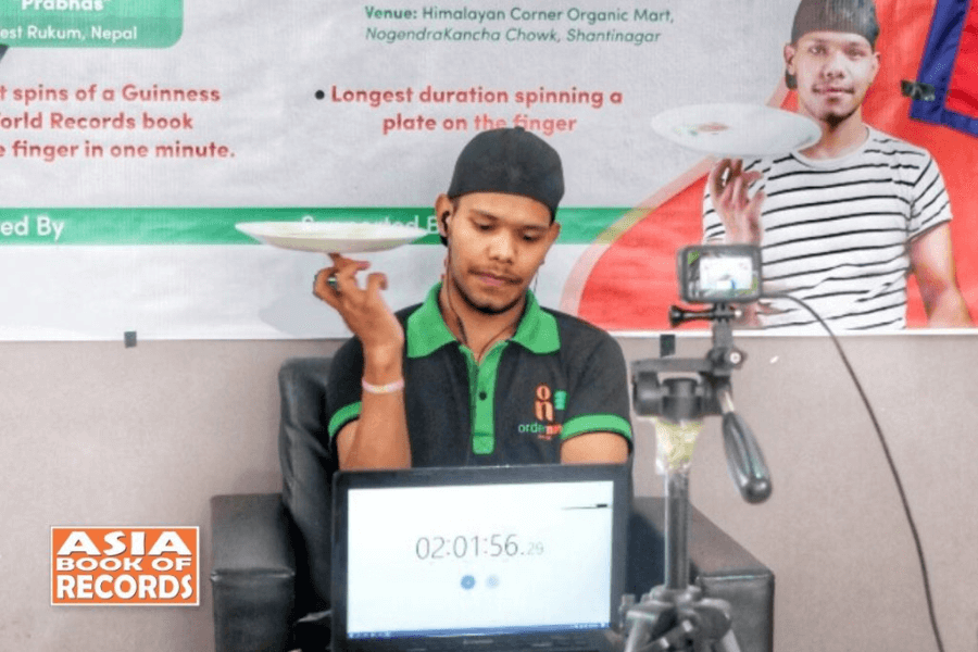 The image shows a man attempting a record for spinning a plate on his finger. He is seated, focused, and dressed in a uniform with a logo, possibly representing a sponsor or organization. In the background, a banner describes the record attempt, while a timer displayed on a laptop shows over two hours of spinning. The "Asia Book of Records" logo is visible, indicating the official nature of the attempt, highlighting the endurance and precision required for this challenge.