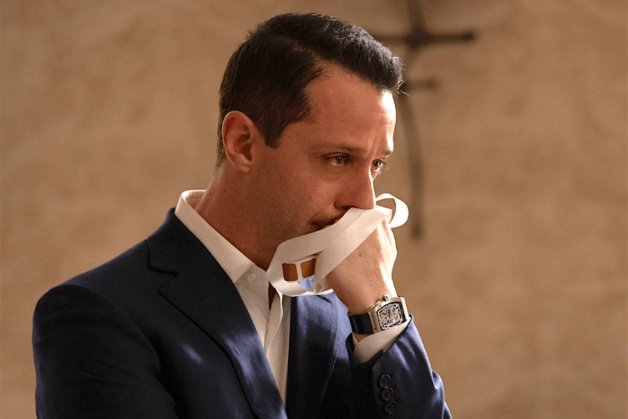 This image shows a man in a formal suit, appearing emotional as he holds a handkerchief to his face. The setting has a muted, warm-toned background with a cross visible, suggesting a solemn or reflective moment. His expression conveys deep introspection or grief.