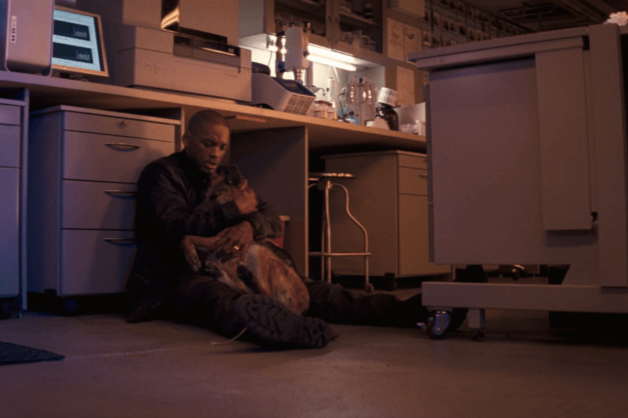 This image depicts a man sitting on the floor in a dimly lit laboratory, holding a German Shepherd dog close to him in an emotional embrace. The setting features desks, drawers, and scientific equipment, illuminated by a mix of warm and cool tones. The man's expression conveys sadness and comfort, highlighting a deep bond with the dog in a moment of vulnerability.