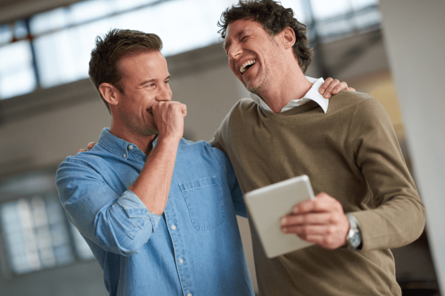 Two men share a hearty laugh, with one holding a tablet and the other covering his mouth in amusement. They are in a bright indoor space with large windows, creating a casual and cheerful atmosphere.