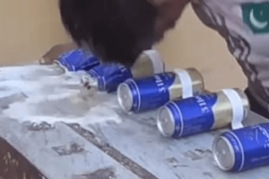 The image shows a person attempting to crush or open a row of blue and gold aluminum cans using their head. The cans are lined up on a rough wooden surface, and liquid is spilling out from one of the crushed cans. The individual appears to be participating in a strength or endurance challenge, possibly part of a record attempt involving unconventional methods for breaking or opening cans. The setting seems informal and outdoors.