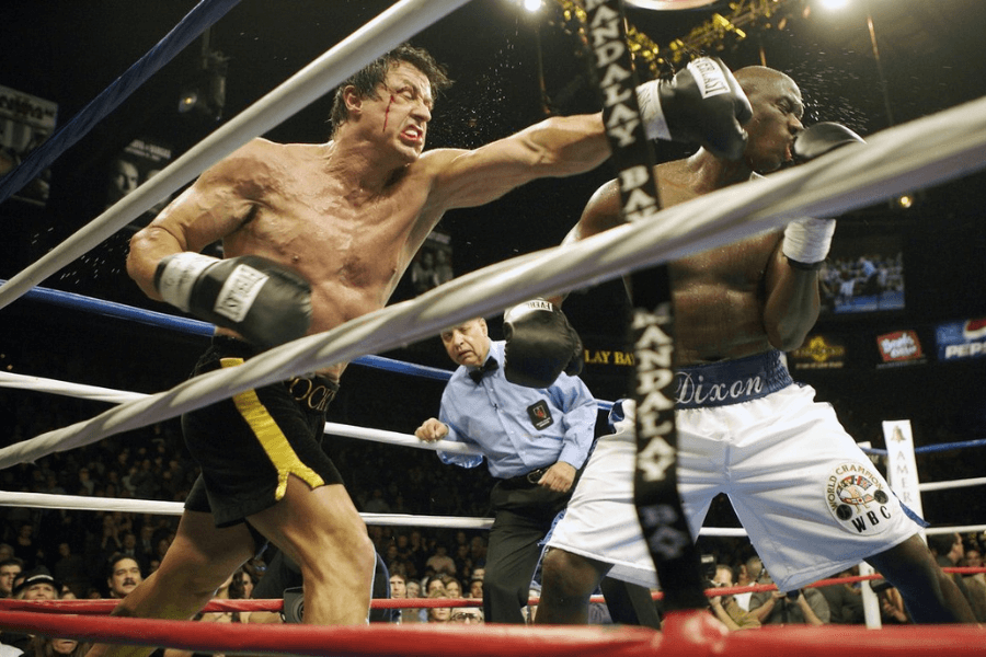 This image depicts an intense boxing match between two fighters in a brightly lit ring, surrounded by an audience. One fighter, shirtless and wearing black shorts with yellow trim, is mid-punch, striking his opponent, who is wearing white shorts. The referee is positioned between them, closely monitoring the action. The scene captures the high energy and drama of a professional fight.