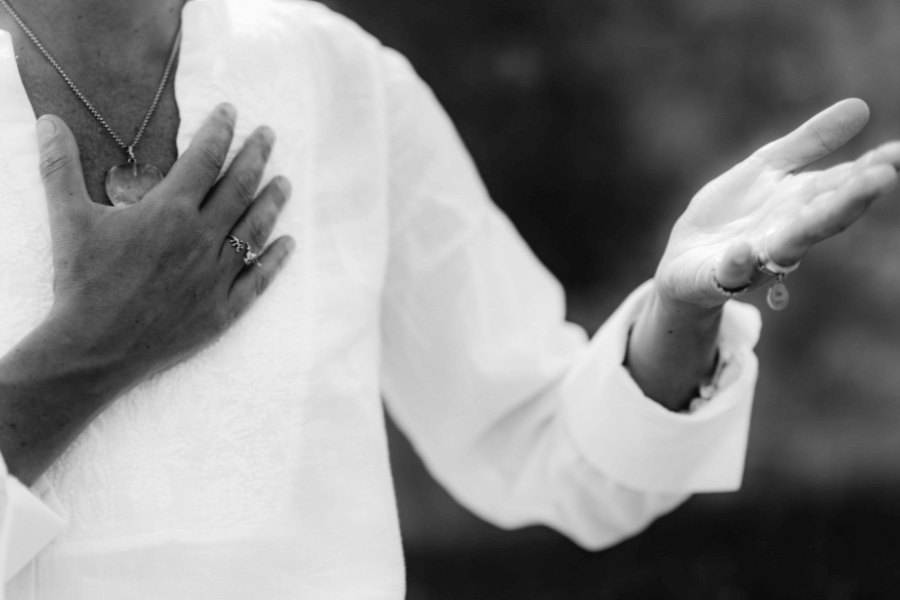 A black-and-white image of a person in a light-colored top with one hand on their chest and the other extended outward in an expressive gesture. The focus is on their hands and jewelry, evoking a sense of sincerity or mindfulness.