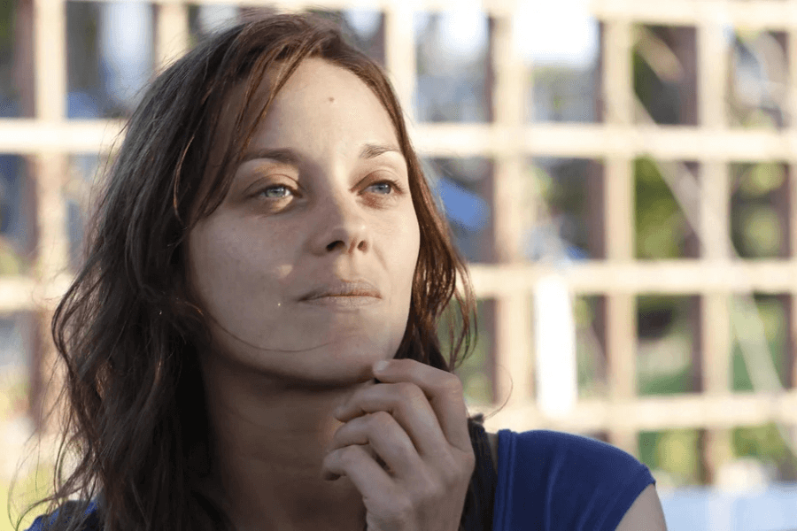 This image shows a woman with a thoughtful expression, gazing upward as she lightly touches her chin. She is bathed in natural sunlight, with a blurred outdoor background featuring a wooden trellis. Her serene demeanor suggests a reflective or peaceful moment.