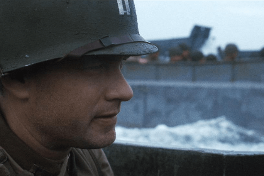 This image shows a soldier wearing a helmet, closely framed in profile, with a serious expression. He is in a military landing craft with turbulent waters visible in the background, along with other soldiers and the steel structure of the boat. The muted color palette and intense atmosphere suggest a tense, war-time setting.