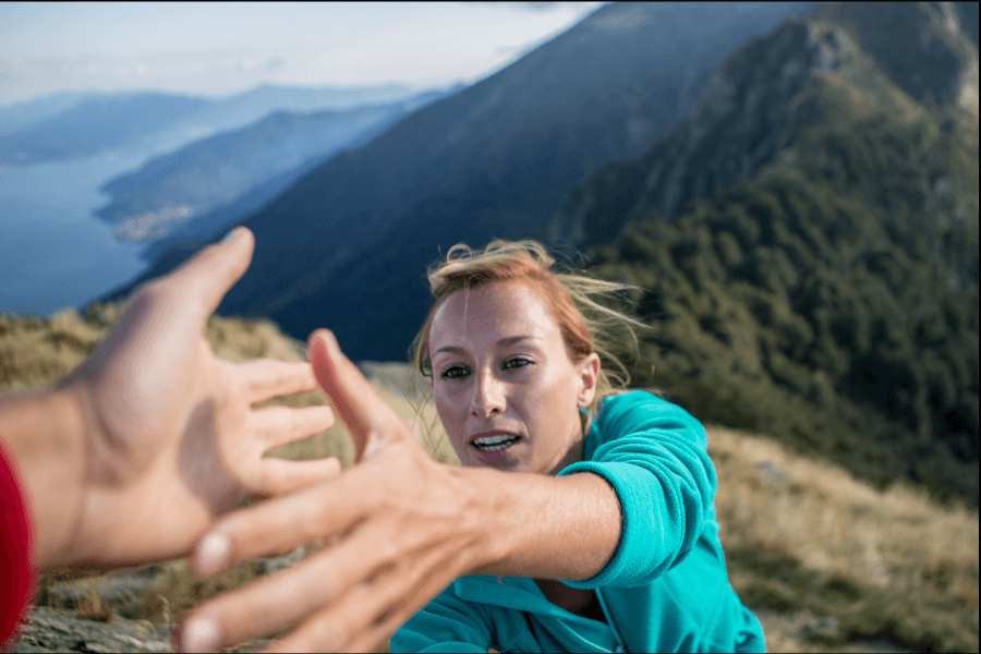 A woman in a turquoise jacket reaches out for help on a steep mountainside, with dramatic landscapes and a distant body of water visible in the background. The image captures a moment of urgency and connection in a rugged outdoor setting.