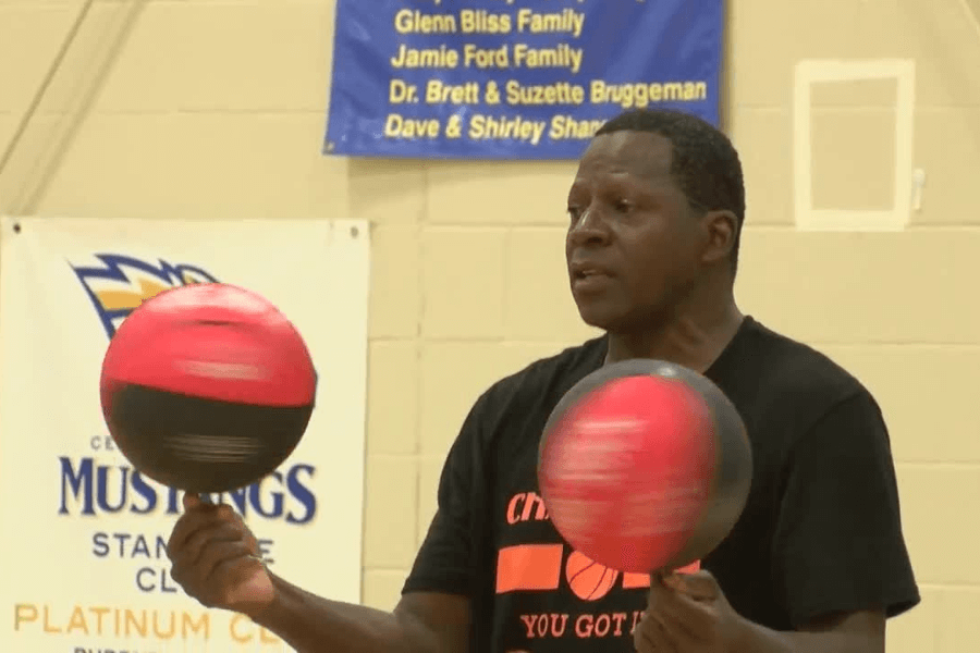 The image shows a man skillfully spinning two basketballs on his fingertips in a gymnasium. He is wearing a black T-shirt with orange text, which appears to be related to basketball or a motivational slogan. The background includes banners and sports equipment, indicating the event is taking place in a formal sports setting. The scene likely captures a demonstration of talent or an attempt at a record for basketball spinning.