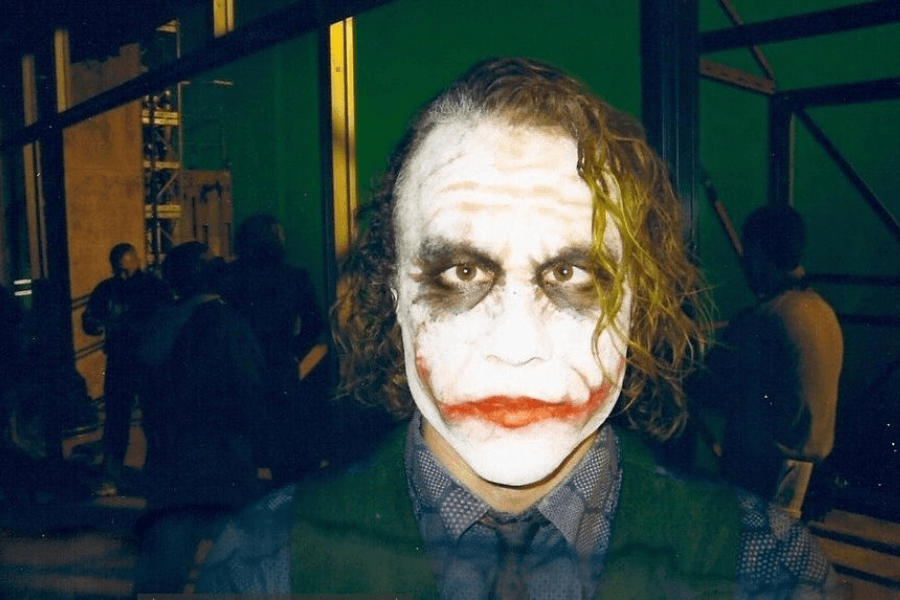 This image shows a character with white face paint, dark smudged eyes, and smeared red lipstick forming an eerie smile. The character has messy, partially green-tinted hair and wears a patterned shirt with a vest, standing in a backstage or studio-like setting.