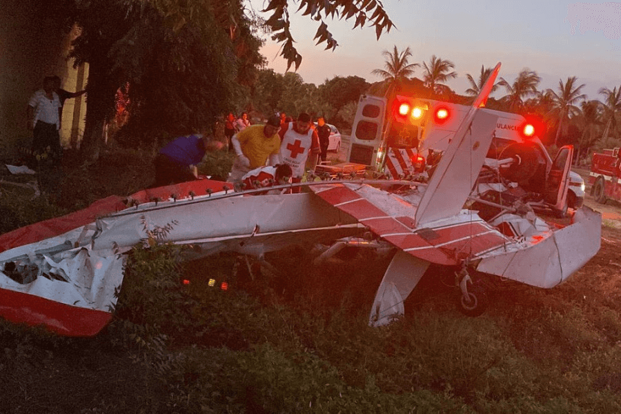 This image shows the wreckage of a small red and white aircraft in a grassy area, surrounded by emergency responders and medical personnel. An ambulance with flashing lights is visible in the background, along with other rescue vehicles, indicating an active response to the incident.