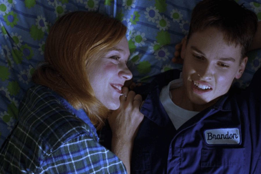 This image shows two individuals lying on a bed with a blue floral-patterned blanket. The woman on the left, wearing a plaid shirt, smiles warmly and leans close to the person on the right, who is dressed in a work uniform with a name tag reading "Brandon." The scene captures an intimate, lighthearted moment between the two.