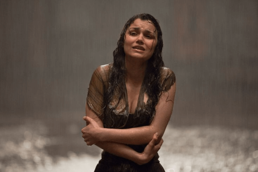 This image shows a woman standing in the rain, soaked and clutching herself with a pained, emotional expression. Her wet hair and the dimly lit, blurred background enhance the sense of vulnerability and despair in the scene. The moment conveys intense emotion, creating a poignant and dramatic atmosphere.