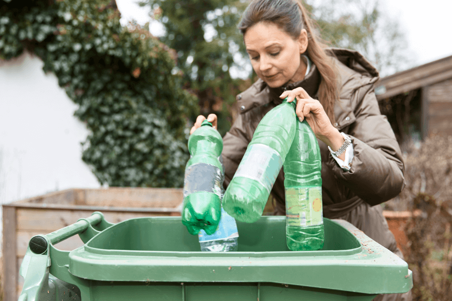 A woman wearing a brown coat recycles plastic bottles by placing them into a green bin. She is outdoors in a garden-like setting, promoting environmental responsibility.