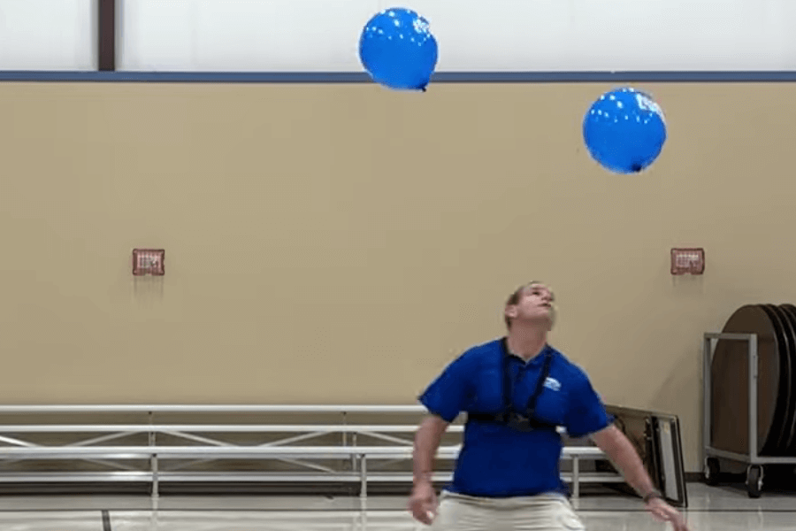 The image shows a man in a blue shirt skillfully keeping two blue balloons afloat in the air using his head. The setting appears to be an indoor gymnasium, with bleachers and folded tables visible in the background. The activity seems to be part of a challenge or record attempt involving balance, coordination, and focus to maintain the balloons in motion without letting them touch the ground.