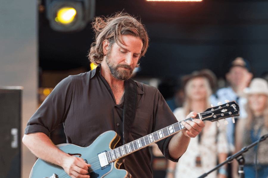 This image shows a man with shoulder-length hair and a beard playing an electric guitar on an outdoor stage. He is dressed in a dark shirt with rolled-up sleeves and appears focused on his performance. The background features blurred onlookers and stage lighting, creating a warm, live music atmosphere.