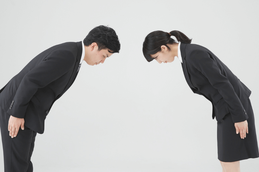 A man and a woman in formal business attire bow to each other in a gesture of respect. The plain white background emphasizes the formality and cultural significance of the interaction.