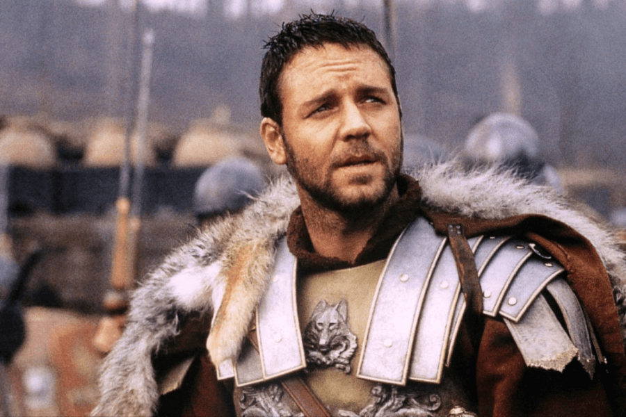 This image depicts a man dressed as a Roman soldier, wearing ornate armor with wolf emblems and a fur-lined cloak. He has a rugged appearance with short, slightly disheveled hair and a beard, gazing upward with a determined expression. The background shows a battlefield setting with blurred soldiers and equipment, creating an epic and historical atmosphere.