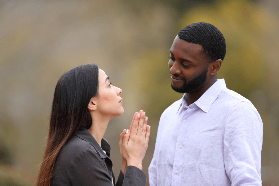 A woman with clasped hands looks up at a man who smiles down at her in an outdoor setting. Their expressions convey a sense of connection and understanding.