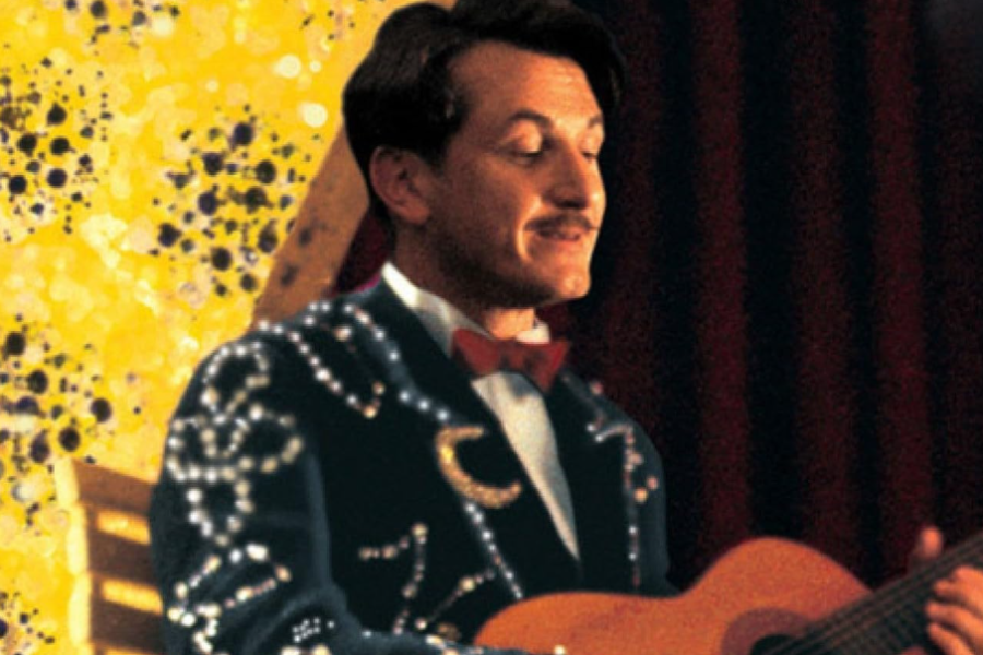 The image shows a man dressed in an ornate, dark suit adorned with sparkling embellishments, including a crescent moon design. He is seated against a bright, decorative yellow background and is playing a guitar, with a subtle, content expression on his face.