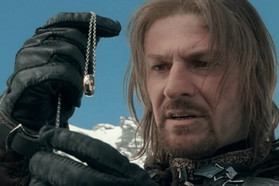 This image shows a man with shoulder-length hair and a solemn expression, holding up a golden ring on a chain in a gloved hand. He appears to be in a snowy, mountainous setting, and his intense gaze conveys a mix of awe and inner conflict. The scene feels dramatic and pivotal, with a focus on the object and its significance.