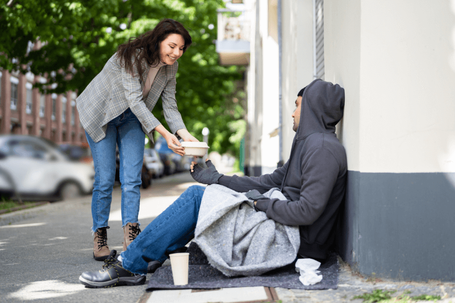 A woman bends down to offer a food container to a man sitting on the sidewalk wrapped in a blanket. The outdoor setting is along a tree-lined street, highlighting an act of kindness and compassion.