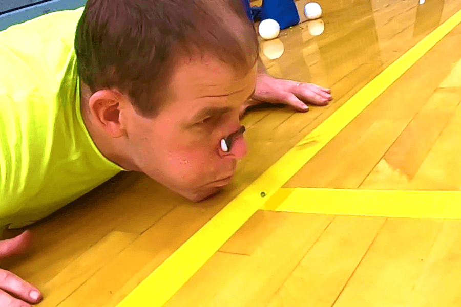 The image shows a man lying on a gym floor, intensely focused on propelling a small object forward with his breath. A ping pong ball appears to be part of the scene, suggesting an unusual competition or record attempt requiring controlled exhalation. The bright yellow floor markings and the reflective surface highlight the unique and quirky nature of this activity.