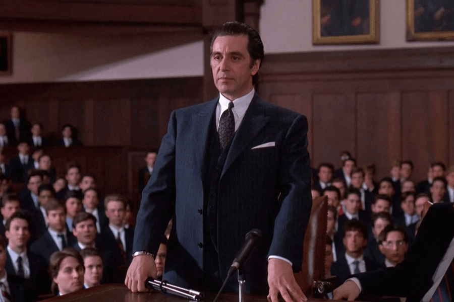 
This image appears to depict a man in a formal suit standing in what looks like a courtroom or a formal assembly setting. He is positioned at the forefront with a serious expression, surrounded by a seated audience dressed in formal attire. 