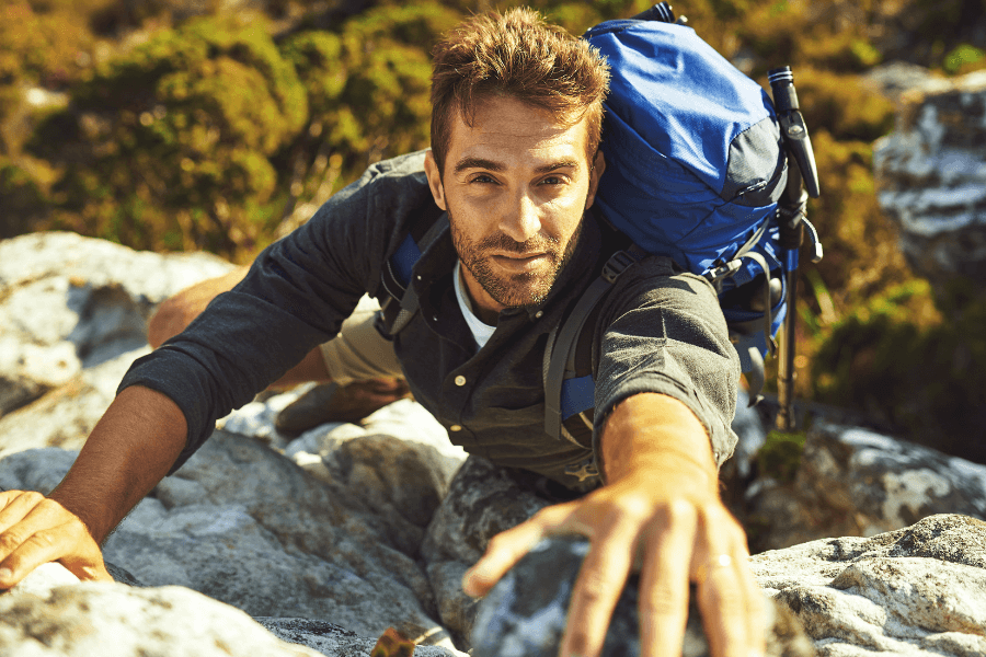 A man with a backpack climbs a rocky terrain, reaching out with determination. The natural backdrop and sunlight emphasize his adventurous spirit.