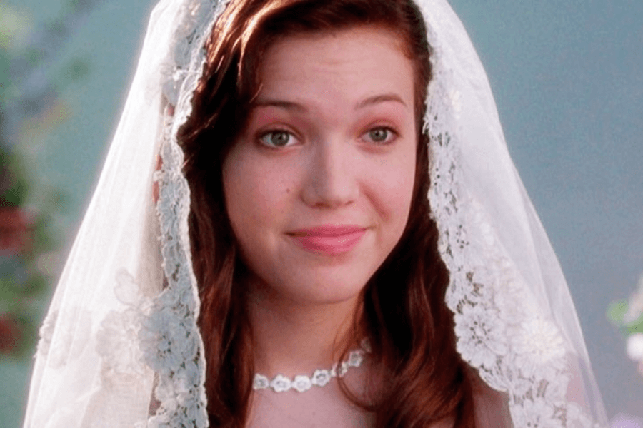 This image shows a young woman with a gentle smile, wearing a delicate lace veil and a simple pearl necklace. The soft, light-colored background enhances the serene and romantic mood, suggesting a wedding or special ceremony. Her expression exudes warmth and happiness.