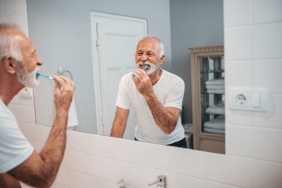 An elderly man cheerfully brushes his teeth while looking into a bathroom mirror. The scene highlights daily self-care and good hygiene habits.