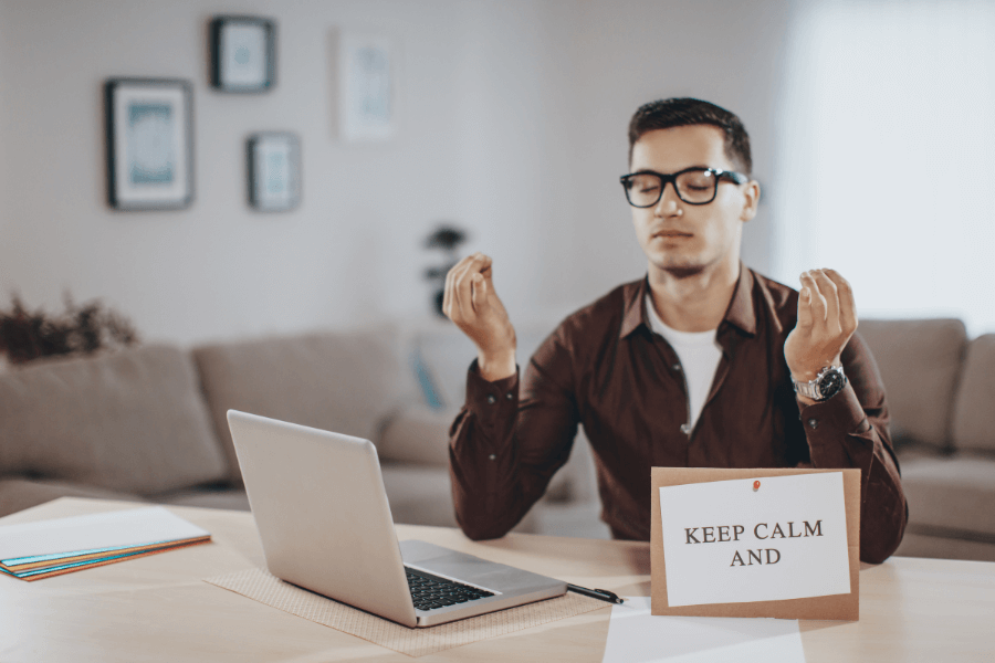 A man wearing glasses sits at a desk with a laptop, holding a meditative pose with closed eyes. A sign on the desk reads "KEEP CALM AND," set against the backdrop of a cozy living room.