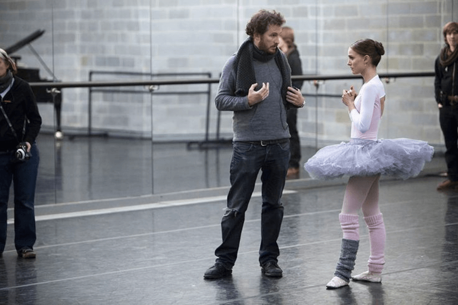 This image shows a ballet studio with a woman dressed in a ballet costume, including a grey tutu and pink leg warmers, speaking to a man wearing casual clothing and a scarf. The setting features mirrored walls and a barre, with additional people and a piano visible in the background.