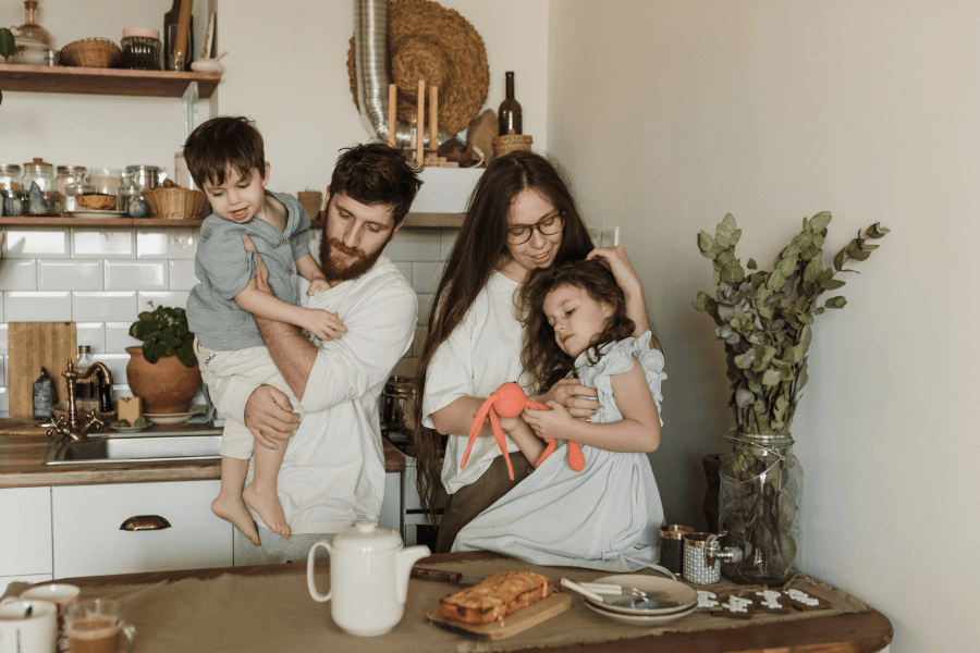 A family in a cozy kitchen, with a father holding a young boy and a mother hugging a little girl holding a stuffed toy. The table in front of them has coffee mugs, a teapot, and a loaf of bread, creating a warm and homey atmosphere.