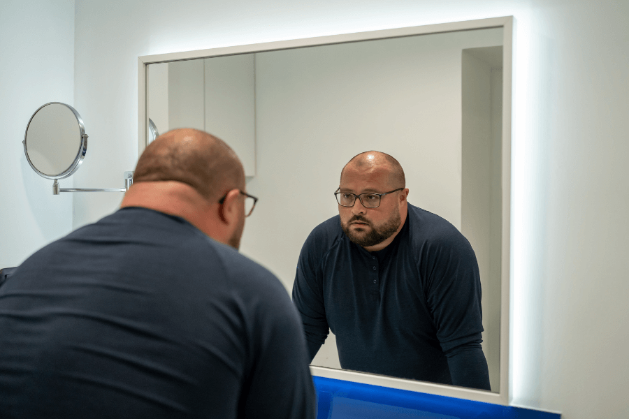 A man wearing glasses and a dark shirt looks at himself intently in a brightly lit bathroom mirror. The room features a minimalist design with a mounted circular mirror on the wall.