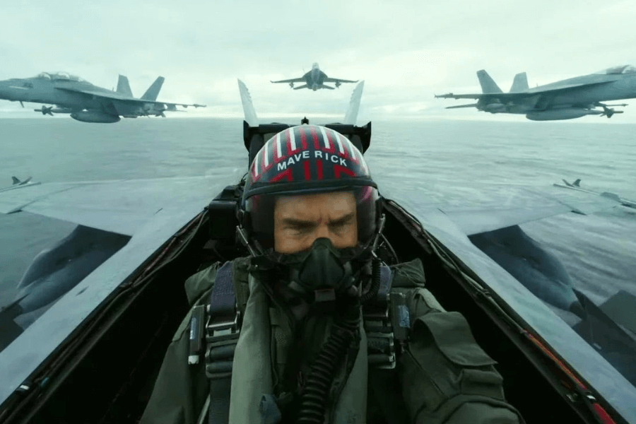 This image shows a pilot in a fighter jet cockpit, wearing a helmet labeled "MAVERICK" and an oxygen mask. The jet is in formation with other aircraft flying over an open body of water, suggesting a high-intensity aviation scenario.