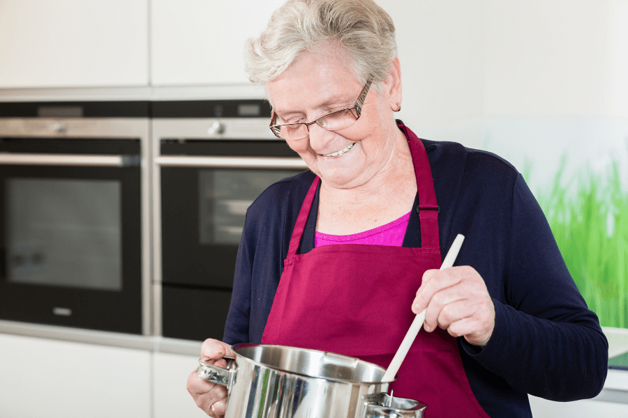 An elderly woman wearing glasses and a maroon apron smiles as she stirs a pot in a modern kitchen. Built-in ovens and a backsplash featuring green grass are visible in the background.