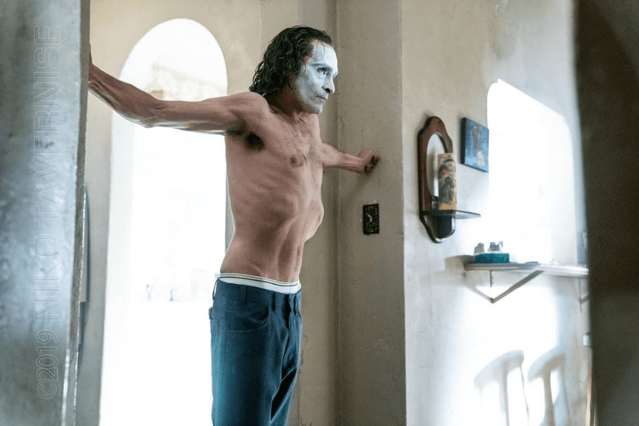 This image depicts a shirtless man with white face paint and green-tinted hair, stretching his arms against a doorway in a dimly lit room. His emaciated frame and intense posture create an unsettling and dramatic atmosphere.