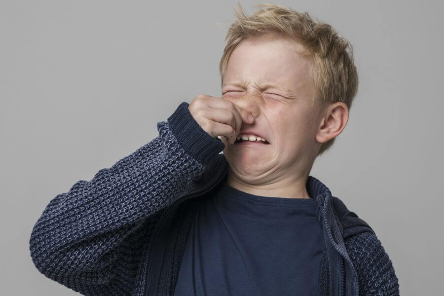 Portrait of boy holding nose while standing against gray background.